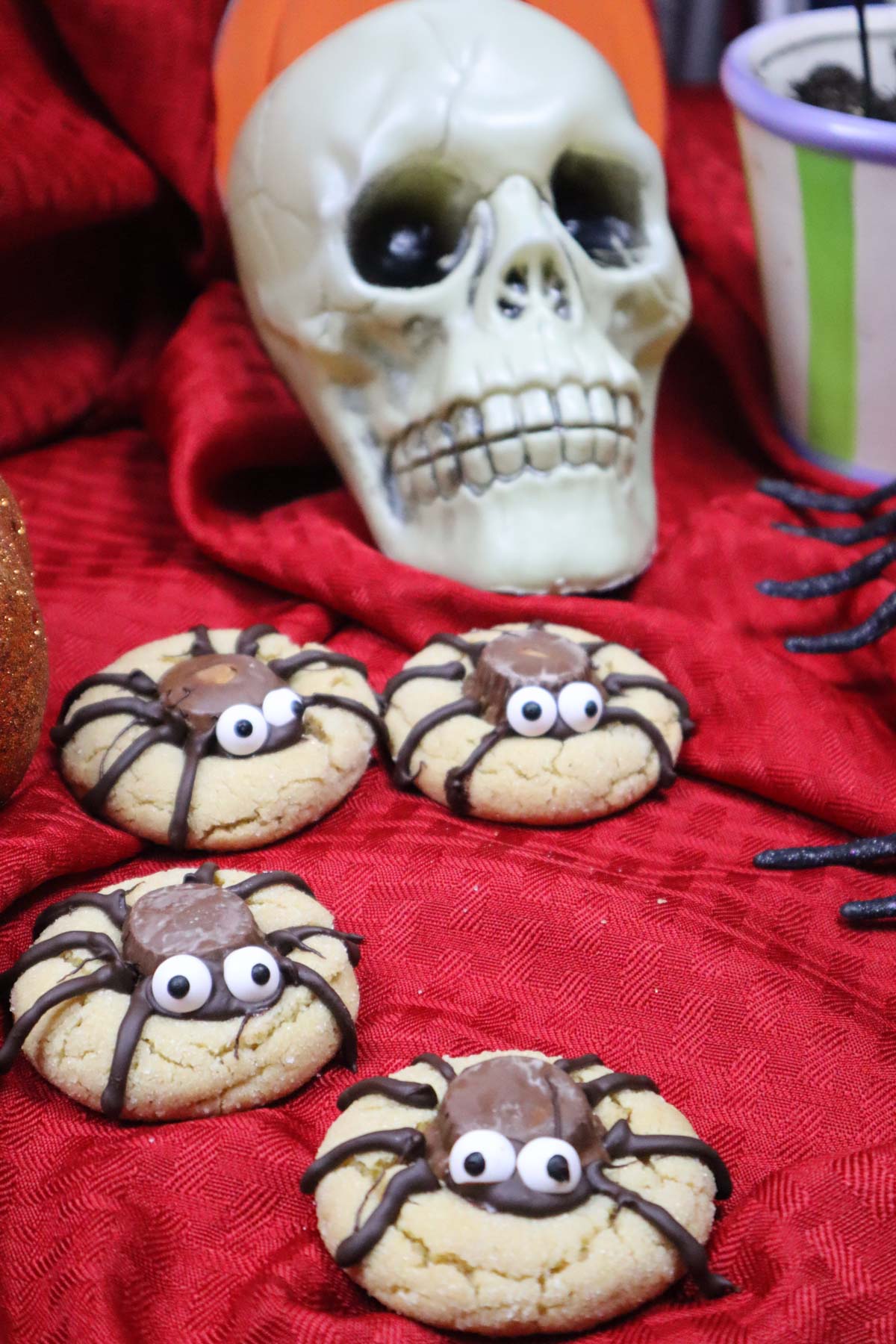 Ragni di Halloween, foto di presentazione con tanti biscotti a forma di ragno su una tovaglia rossa e uno scheletro decorativo di halloween sullo sfondo.